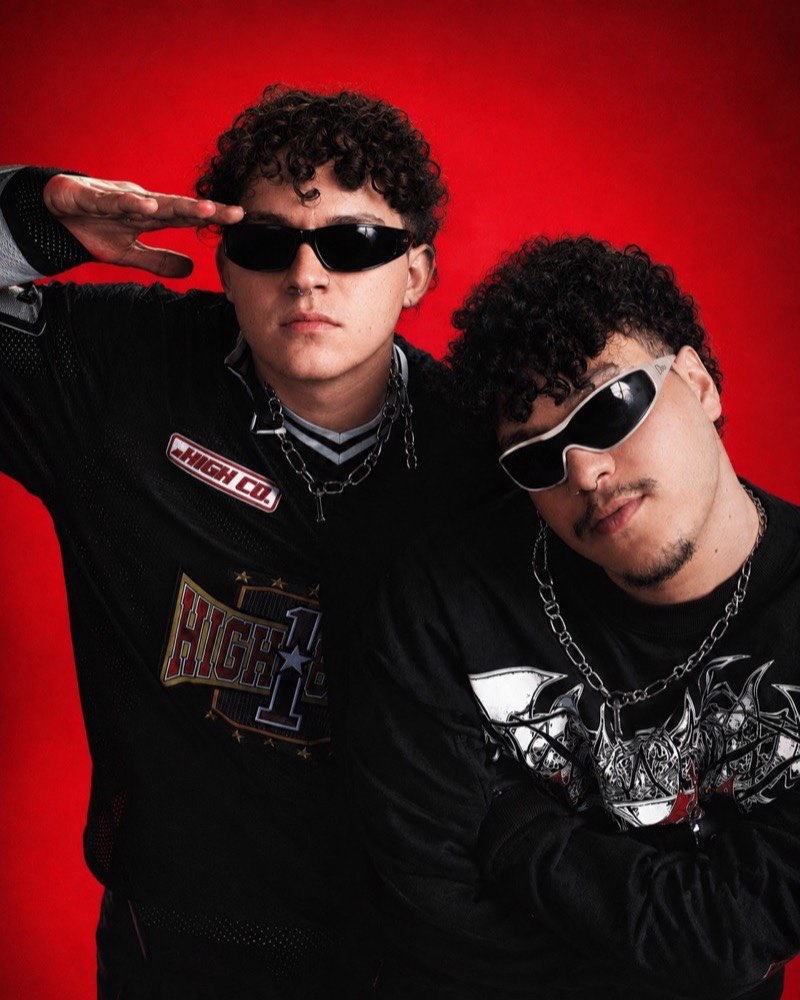 A dupla da somma posando sobre fundo vermelho, de óculos escuros e roupas pretas.