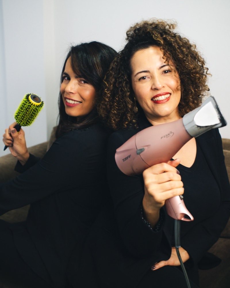 As duas profissionais do NP Studio Hair posando de costas uma para a outra, com escova e secador na mão.